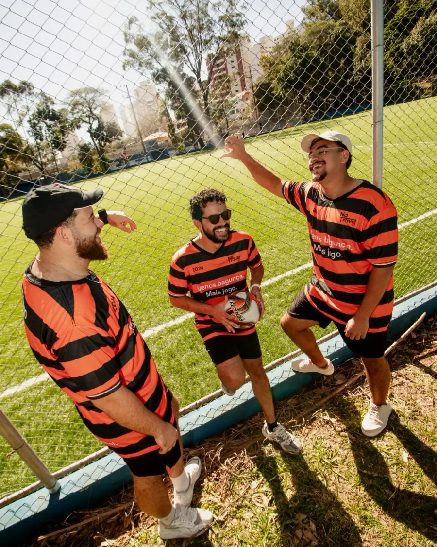 Três integrantes com camisetas laranjas e pretas do NaTrave! conversam junto à grade do campo